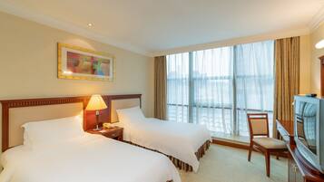 Ein Hotelzimmer mit zwei Betten, einem Schreibtisch mit Lampe, einem Fernseher und einem großen Fenster mit Vorhängen.