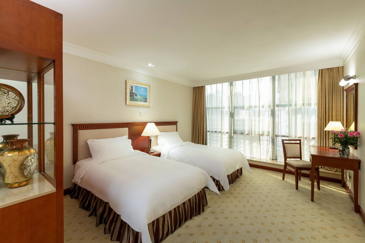 A hotel room with two beds, a desk, a chair, a vase with flowers, and a large window with curtains.