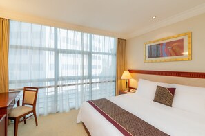 Habitación de hotel con una cama grande, un escritorio y una silla. Se ve una ventana con cortinas translúcidas, un cuadro en la pared y una lámpara en la mesita de noche.