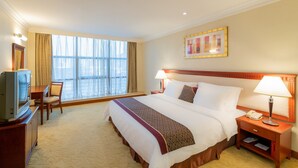 A hotel room with a large bed, a desk with a television, a nightstand with a lamp, and a window with curtains.