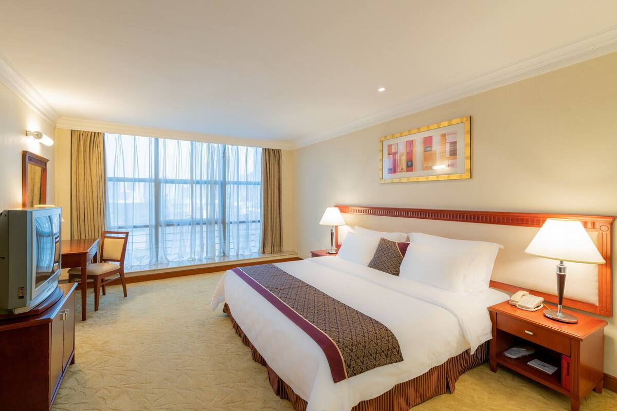 A hotel room with a large bed, a desk with a television, a nightstand with a lamp, and a window with curtains.