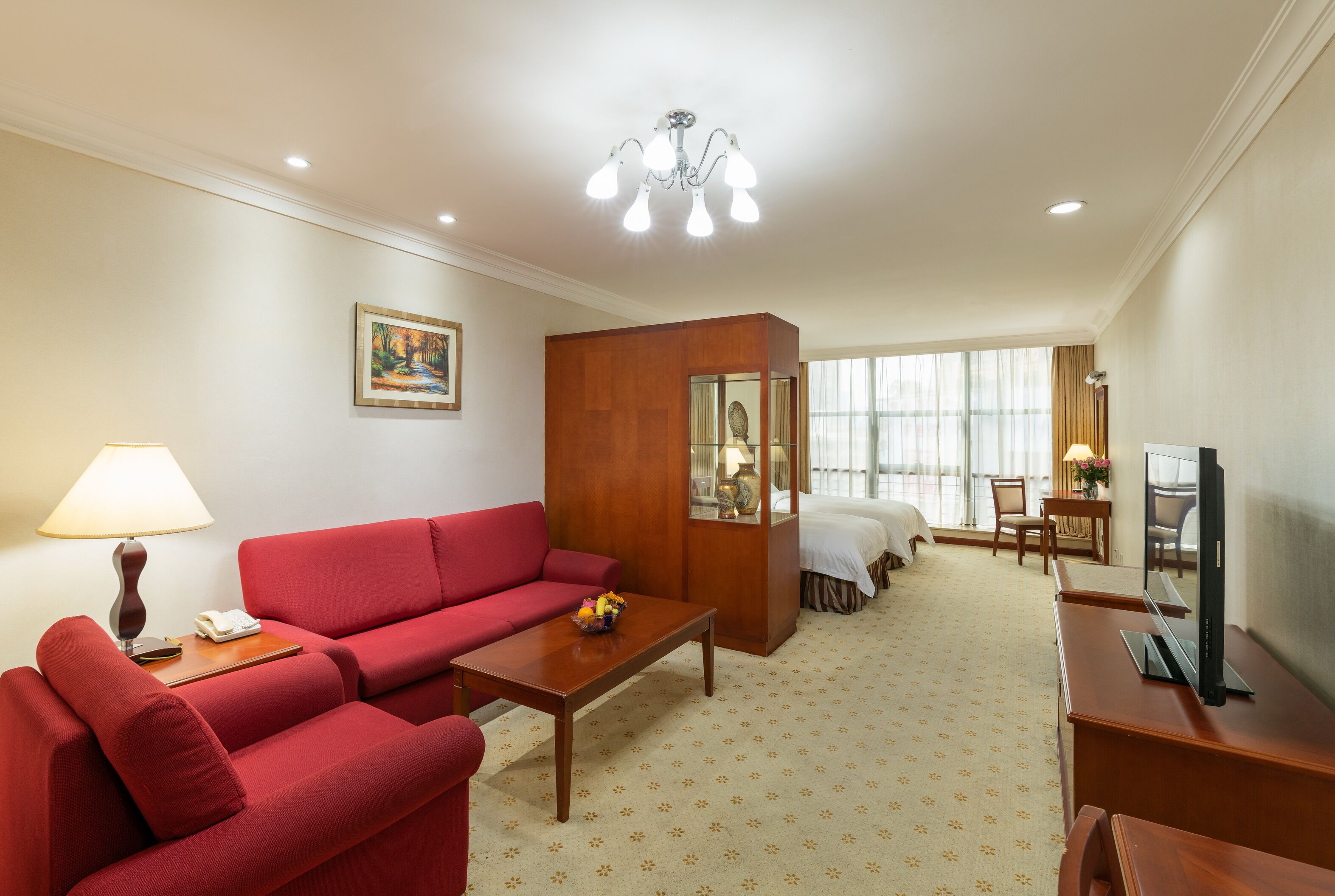 A hotel room with a red sofa, a wooden coffee table, a TV, a desk, and a large window with curtains.