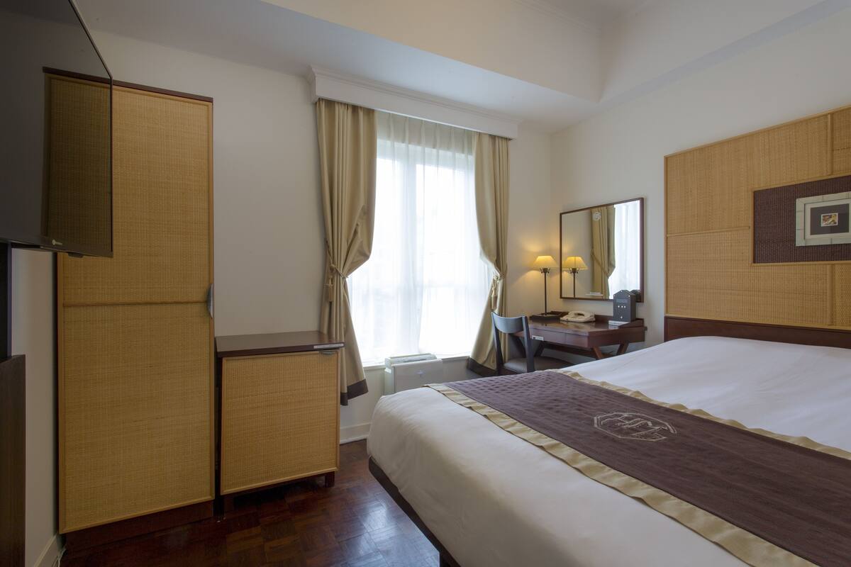 A hotel room with a bed, a desk, a chair, a wardrobe, and a window with curtains.