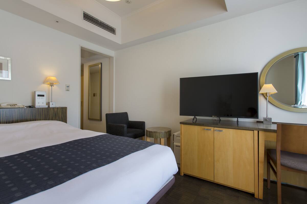 A hotel room with a large bed, a desk with a flat-screen TV, a chair, and a round mirror on the wall.