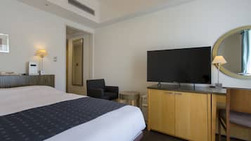 A hotel room with a large bed, a desk with a flat-screen TV, a chair, and a round mirror on the wall.