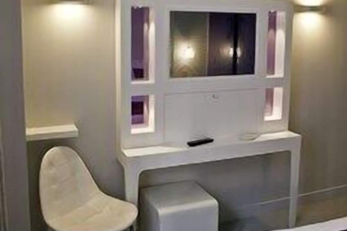 A bedroom with a bed, a white chair, a TV mounted on the wall, and two wall-mounted lights.
