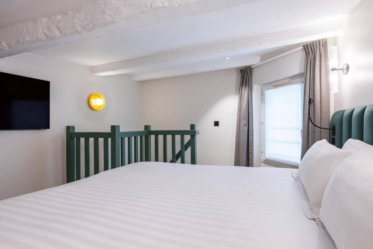 A hotel room with a bed, a television mounted on the wall, a window with curtains, and a bedside lamp.