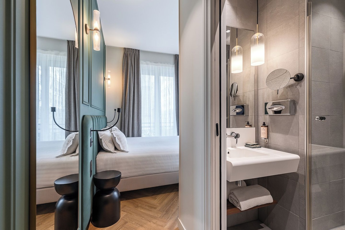 A modern hotel room with a bed, bedside table, and a bathroom with a sink and mirror.