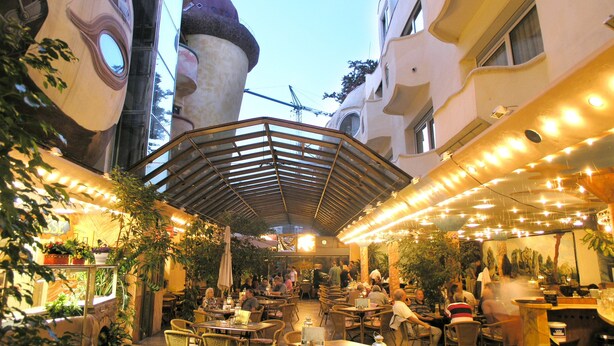 Un área de comedor al aire libre con mesas y sillas, rodeada de edificios de arquitectura singular.