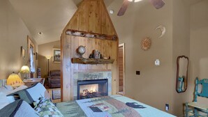 Un dormitorio acogedor con chimenea, una cama con manta estampada, techo de madera y un espejo montado en la pared.