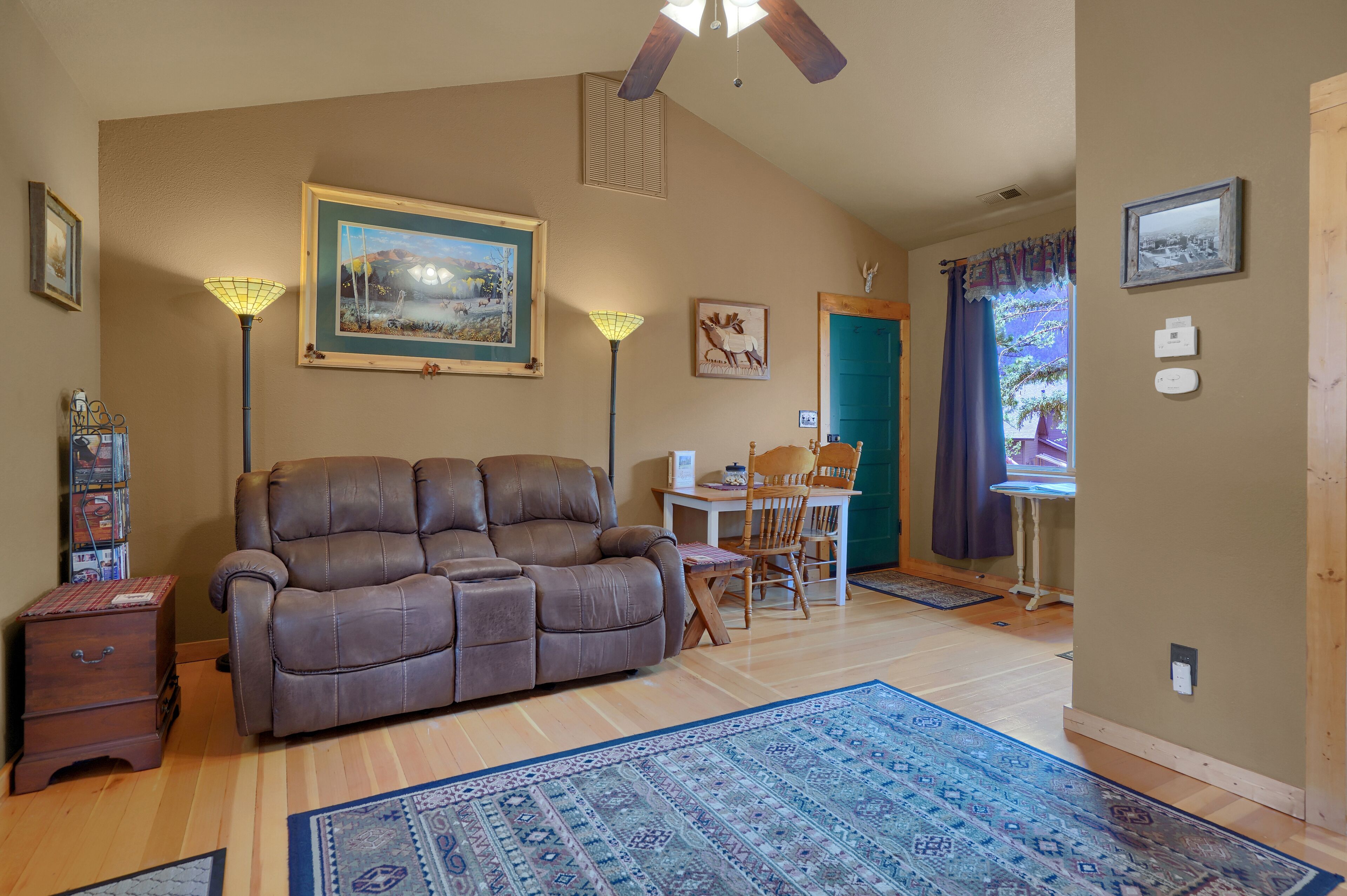 A living room with a brown leather sofa, a dining table with chairs, a blue patterned rug, and a ceiling fan.