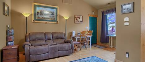 A living room with a brown leather sofa, a dining table with chairs, a blue patterned rug, and a ceiling fan.