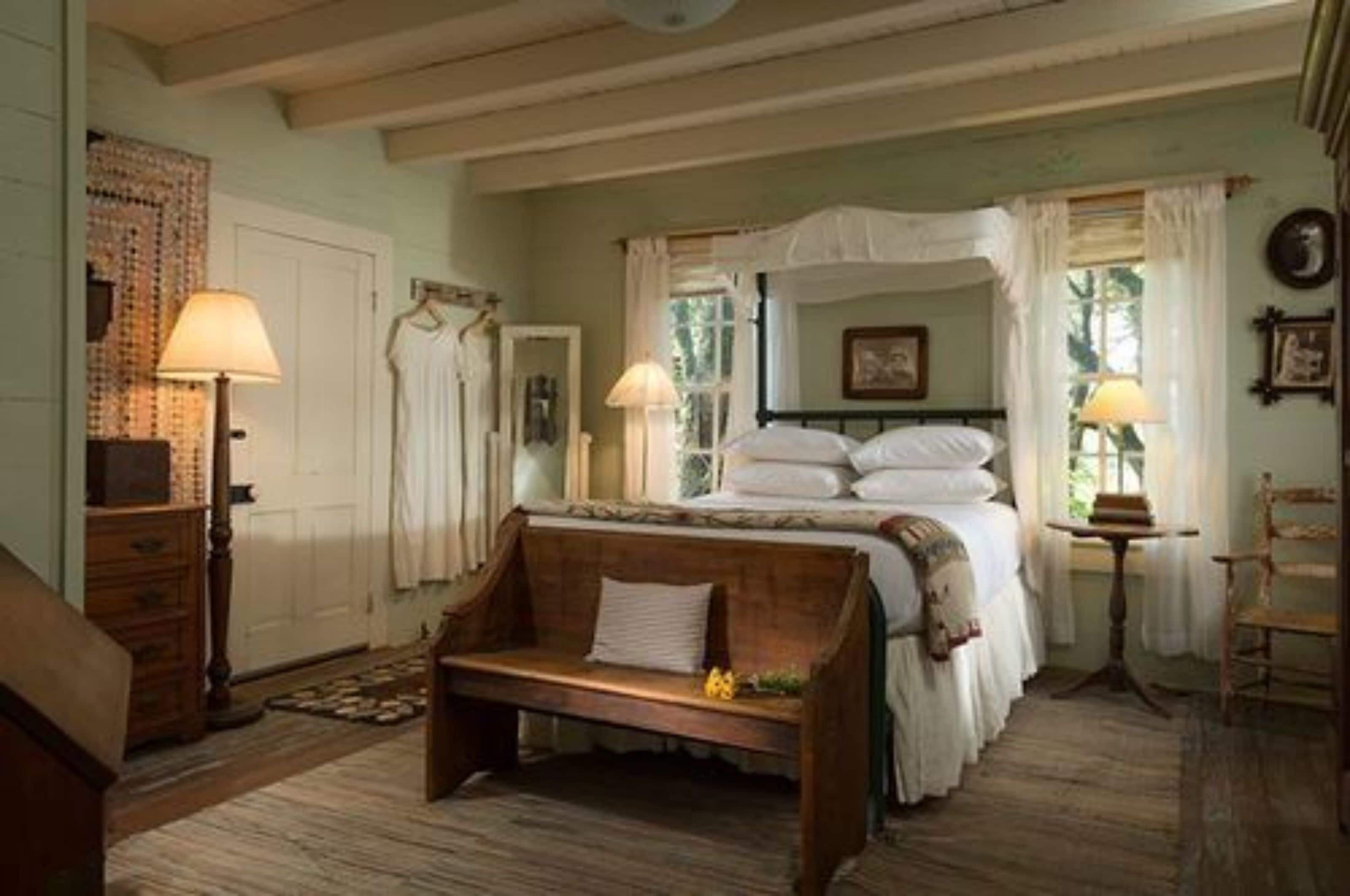 A bedroom with a four-poster bed, two bedside lamps, a wooden bench, and a small table with a book.