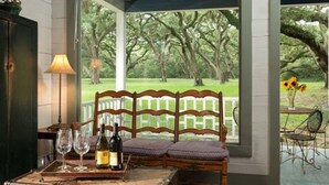 A covered patio with a wooden bench, a table with a bottle and glasses, and a view of a green landscape with trees.