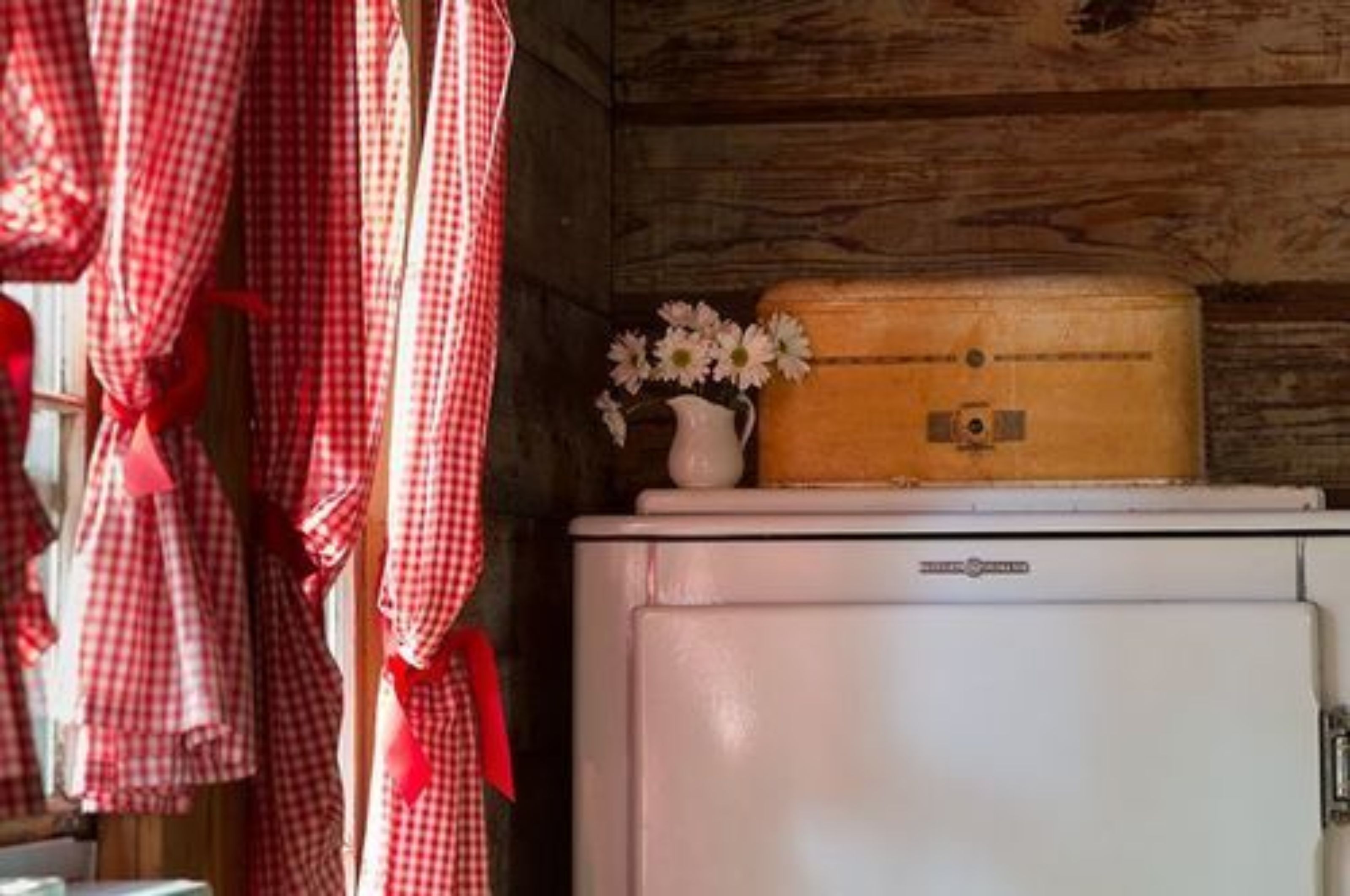 A vintage refrigerator with a wooden case on top, a white pitcher with flowers, and red checkered curtains.