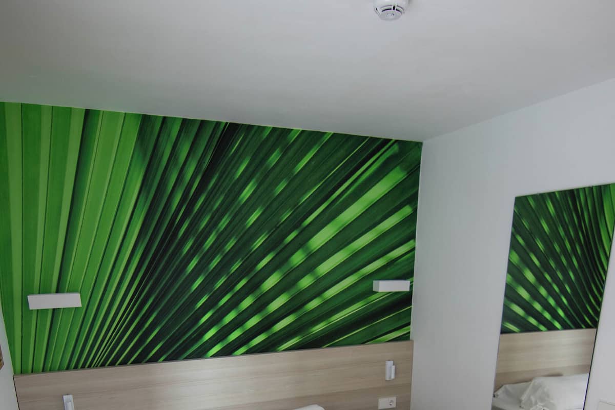 A hotel room with a bed, a green wall with leaf patterns, a mirror, and a green stool.