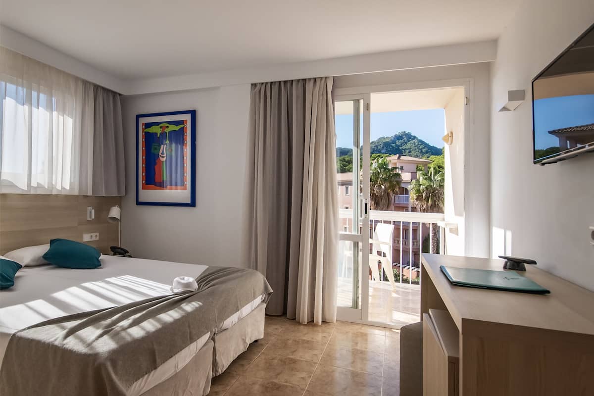 A hotel room with a bed, a desk, a laptop, a painting, and a view of the outside.