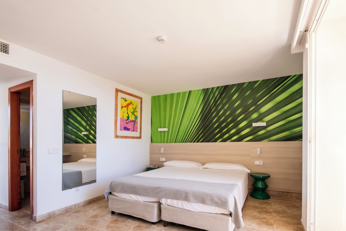 A bedroom with a bed, a mirror, a green leaf-patterned wall, and a small green side table.