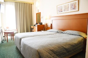Quarto de hotel com duas camas, uma escrivaninha, uma cadeira, uma luminária e um quadro na parede.