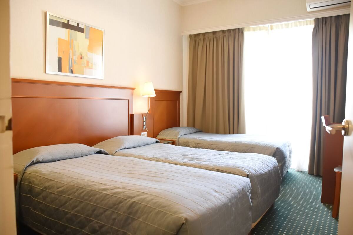 A hotel room with two beds, a nightstand with a lamp, a painting on the wall, and a window with curtains.