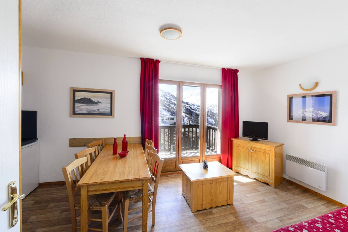 A room with a wooden dining table, chairs, a small wooden cabinet, a television, and a window with red curtains.