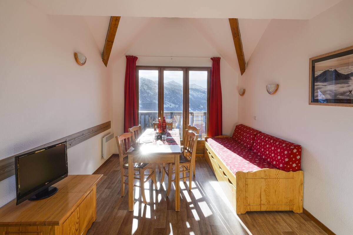 A room with a wooden table, chairs, a TV, a sofa, and a window with a mountain view.