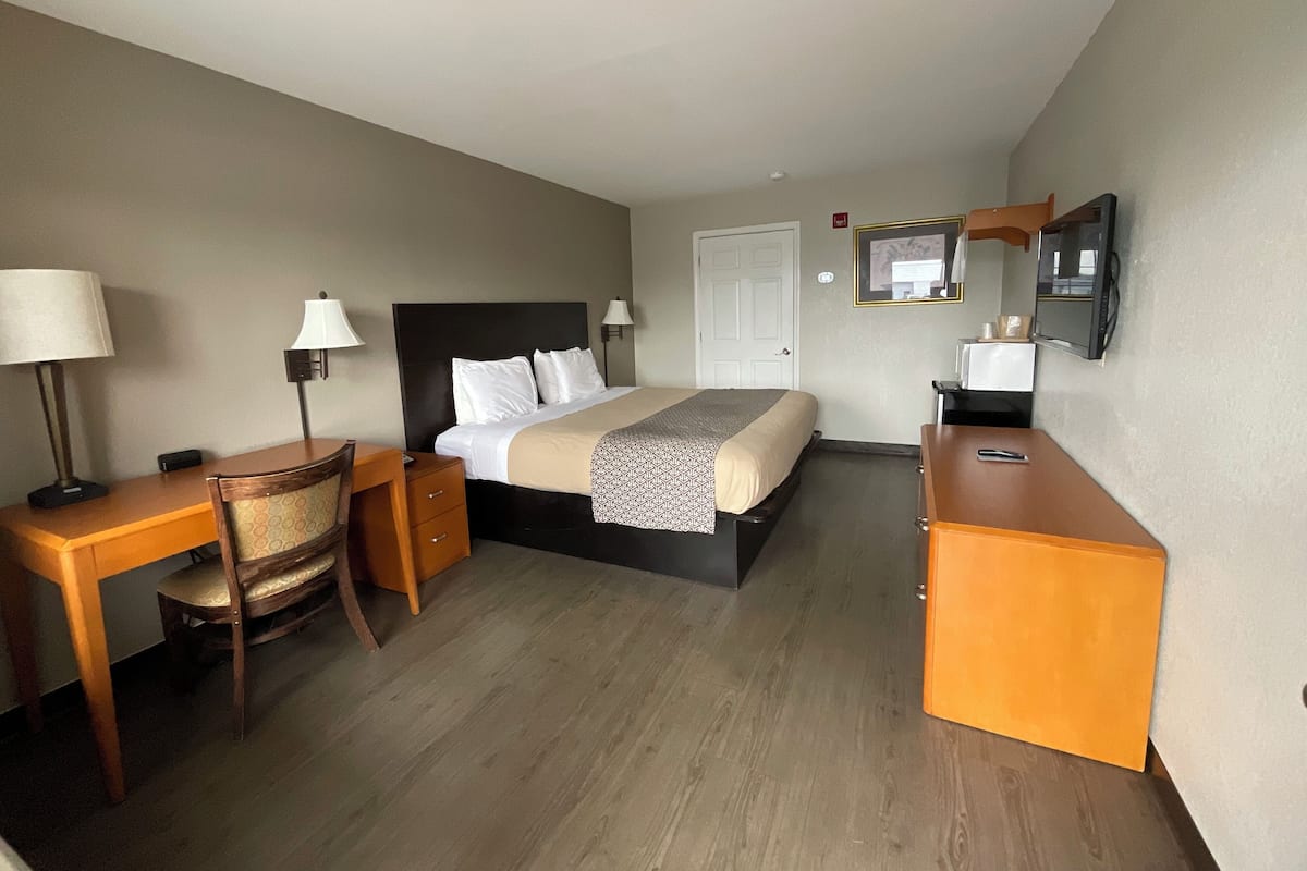 A hotel room with a bed, desk, chair, bedside lamps, a dresser, a TV, and a door.