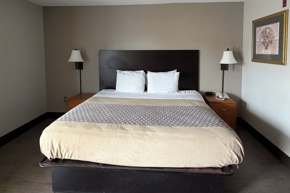 A hotel room with a bed, two bedside tables, two lamps, a framed picture on the wall, and a wooden floor.