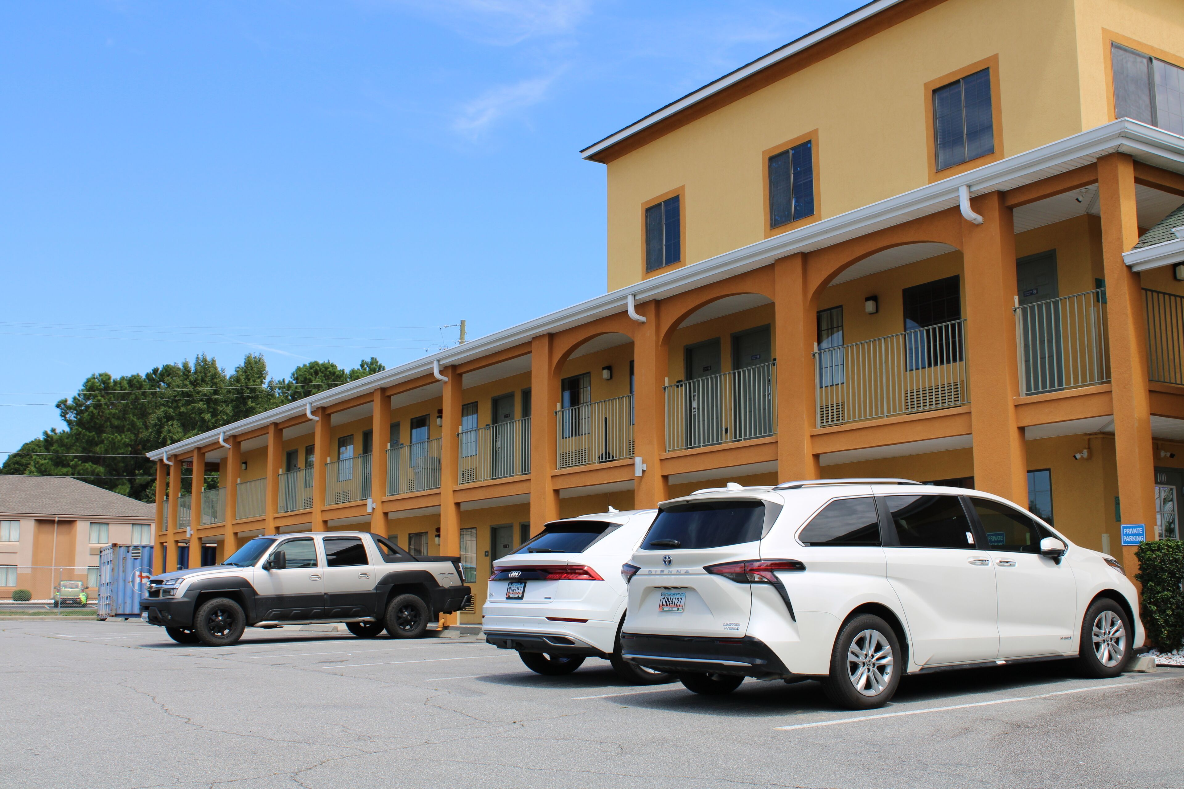 Un hôtel à étages, doté de balcons et d’un parking où sont stationnés plusieurs véhicules.