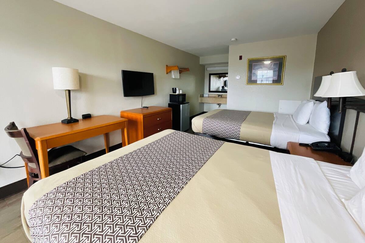 A hotel room with a bed, a desk, a chair, a TV, a mirror, and a bedside table with a lamp.
