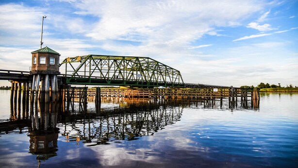 A large green metal bridge over calm water with a small tower on the left.