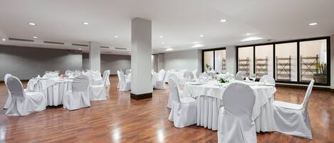 A spacious banquet hall with multiple round tables and white chairs, large windows, and a polished wooden floor.