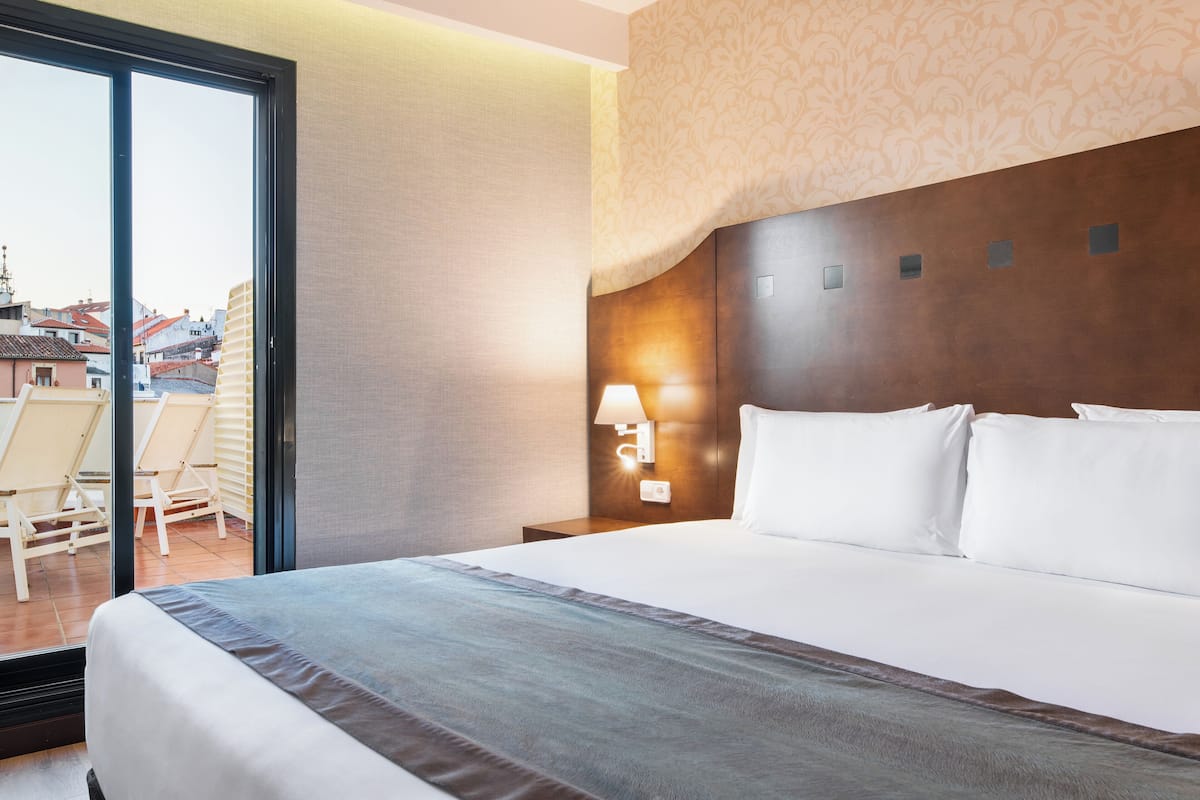 A hotel room with a large bed, a nightstand with a lamp, a view of the city through a sliding door, and a balcony with chairs.