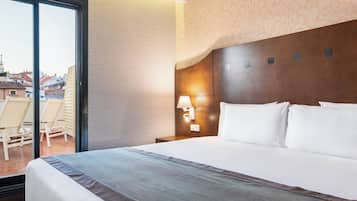 Habitación de hotel con una cama grande, una mesita de noche con lámpara, vista de la ciudad a través de una puerta corrediza y un balcón con sillas.