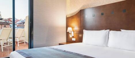 A hotel room with a large bed, a nightstand with a lamp, a view of the city through a sliding door, and a balcony with chairs.