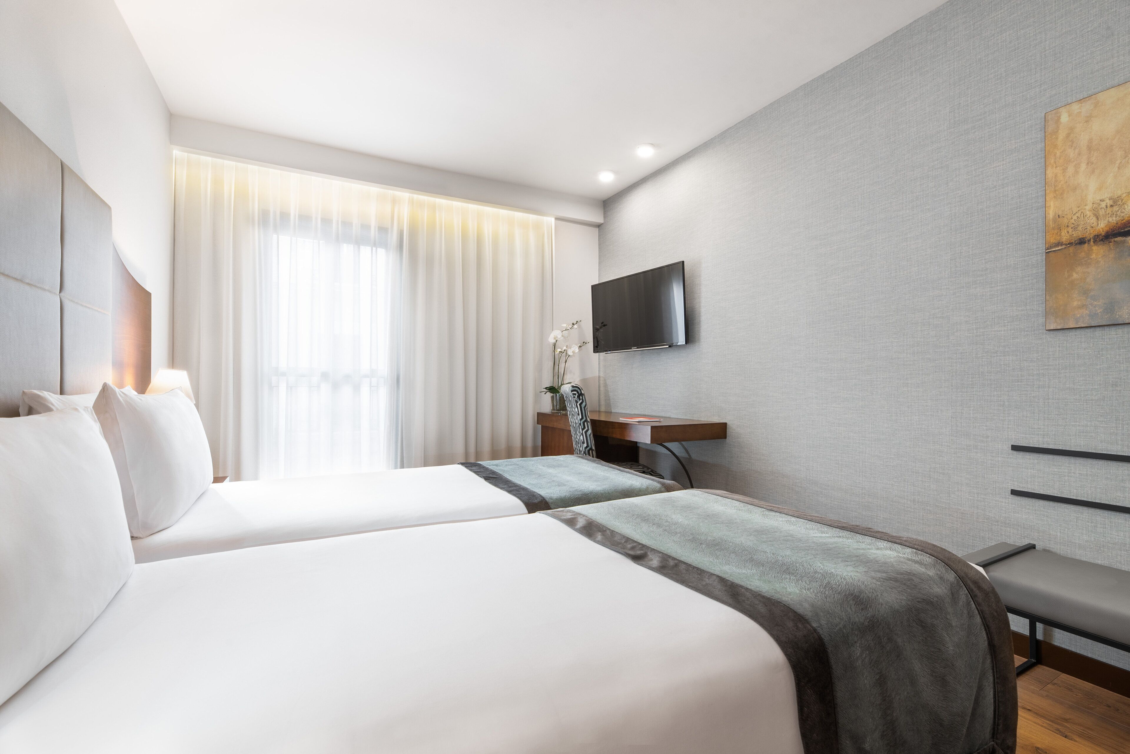 Habitació d'hotel moderna amb dos llits, un televisió de pantalla plana penjada a la paret i un gran finestral amb cortines translúcides.