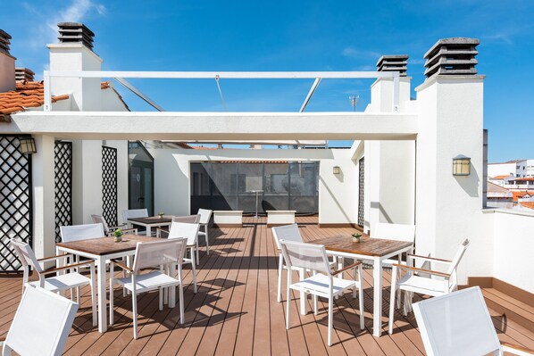 An outdoor patio with white tables and chairs, a wooden floor, and a view of the cityscape. - Dorma Ágora Caceres (Cáceres)