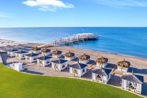 A beachside resort with thatched-roof cabanas, a pier extending into the sea, and a clear blue sky.