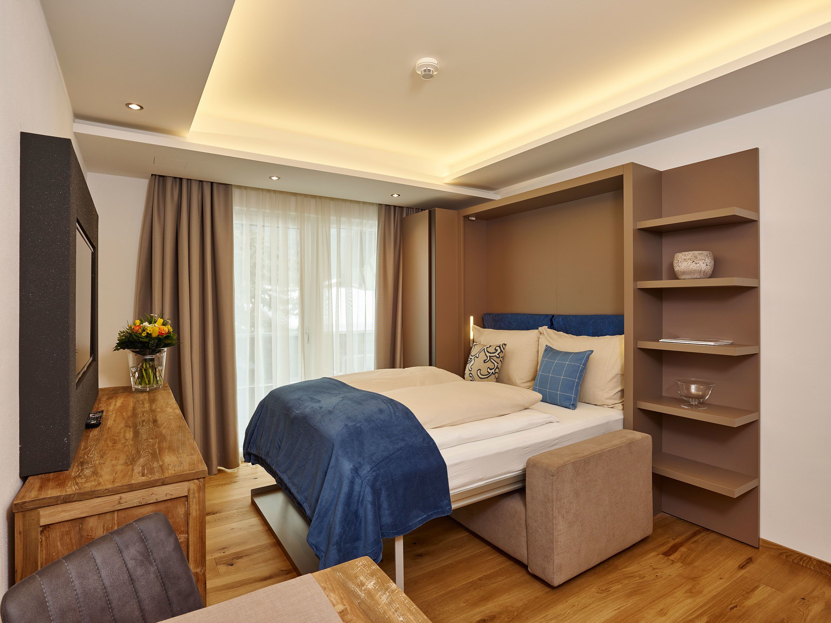 A modern hotel room with a large bed, bedside tables, a wooden desk, and a built-in shelving unit.