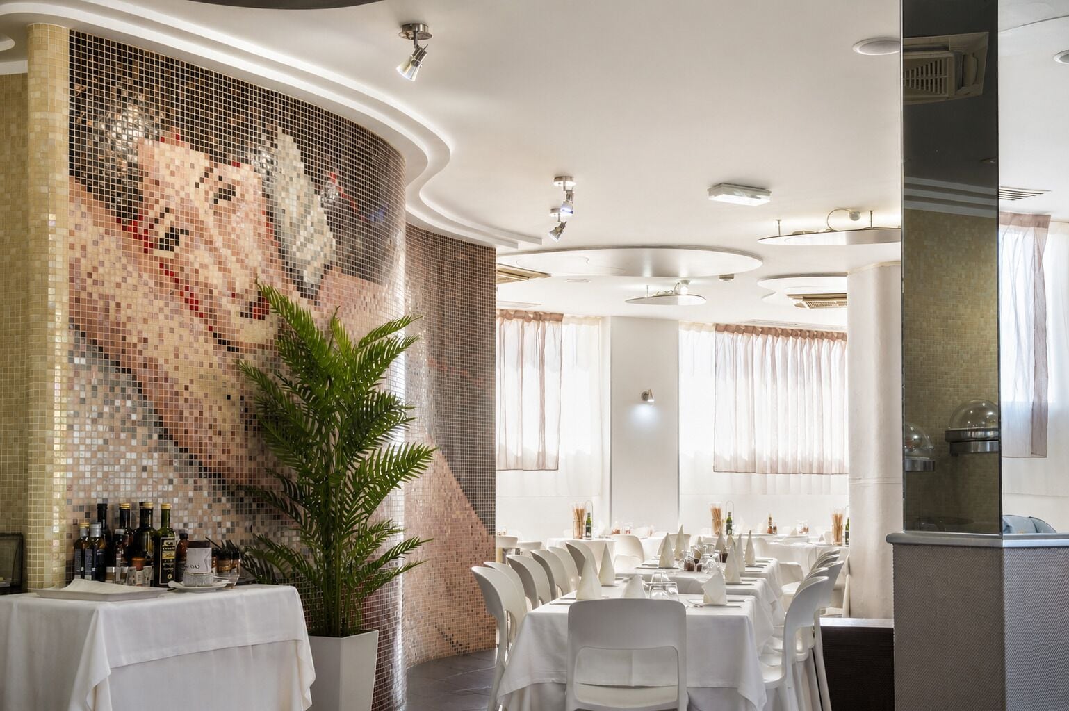 A restaurant with white tablecloths, a mosaic wall, and a potted plant.