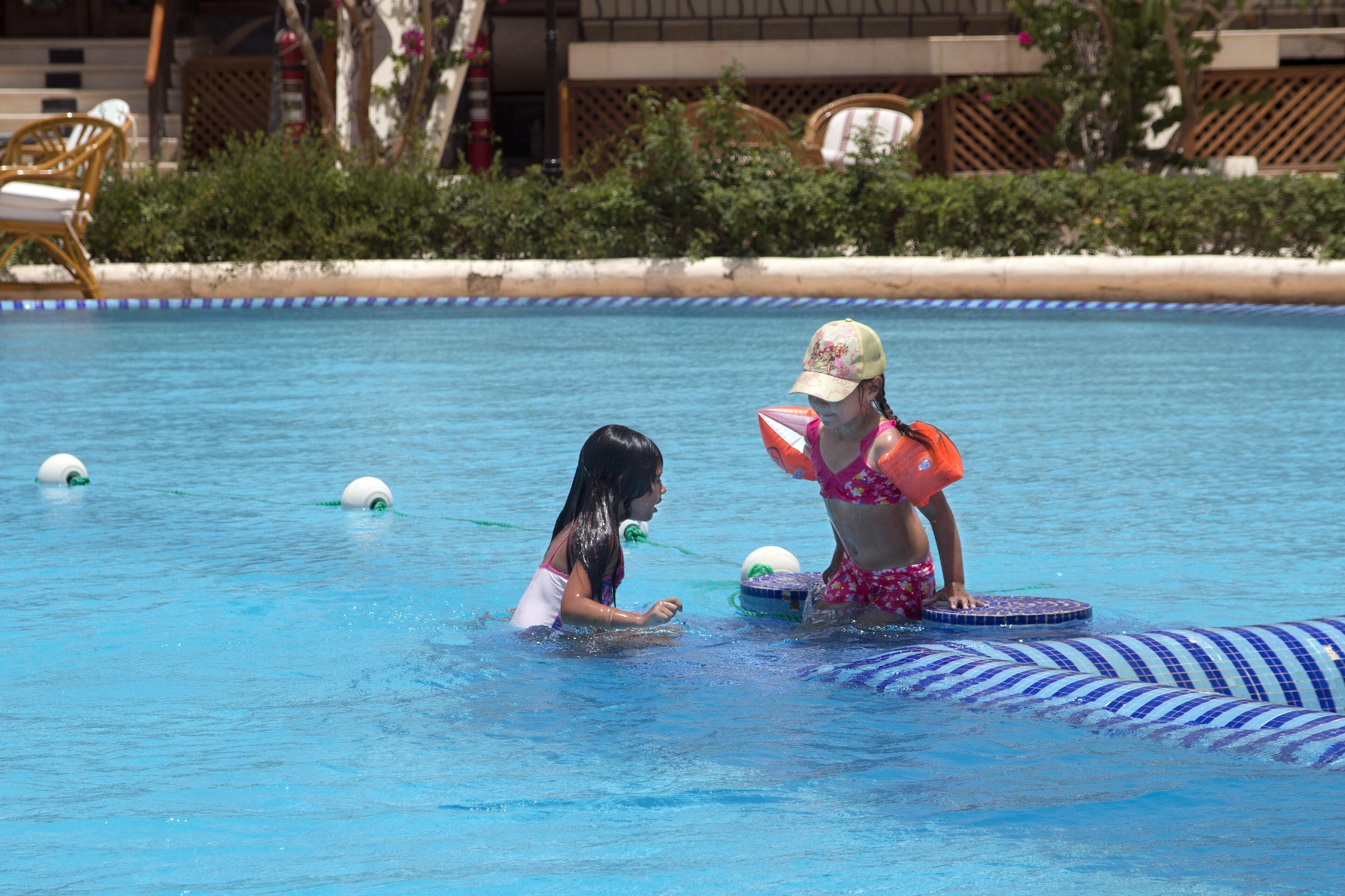 Una piscina con due persone a nuoto, una con costume floreale e l'altra con un costume rosa. Sullo sfondo, sedie a sdraio e un edificio con un balcone.