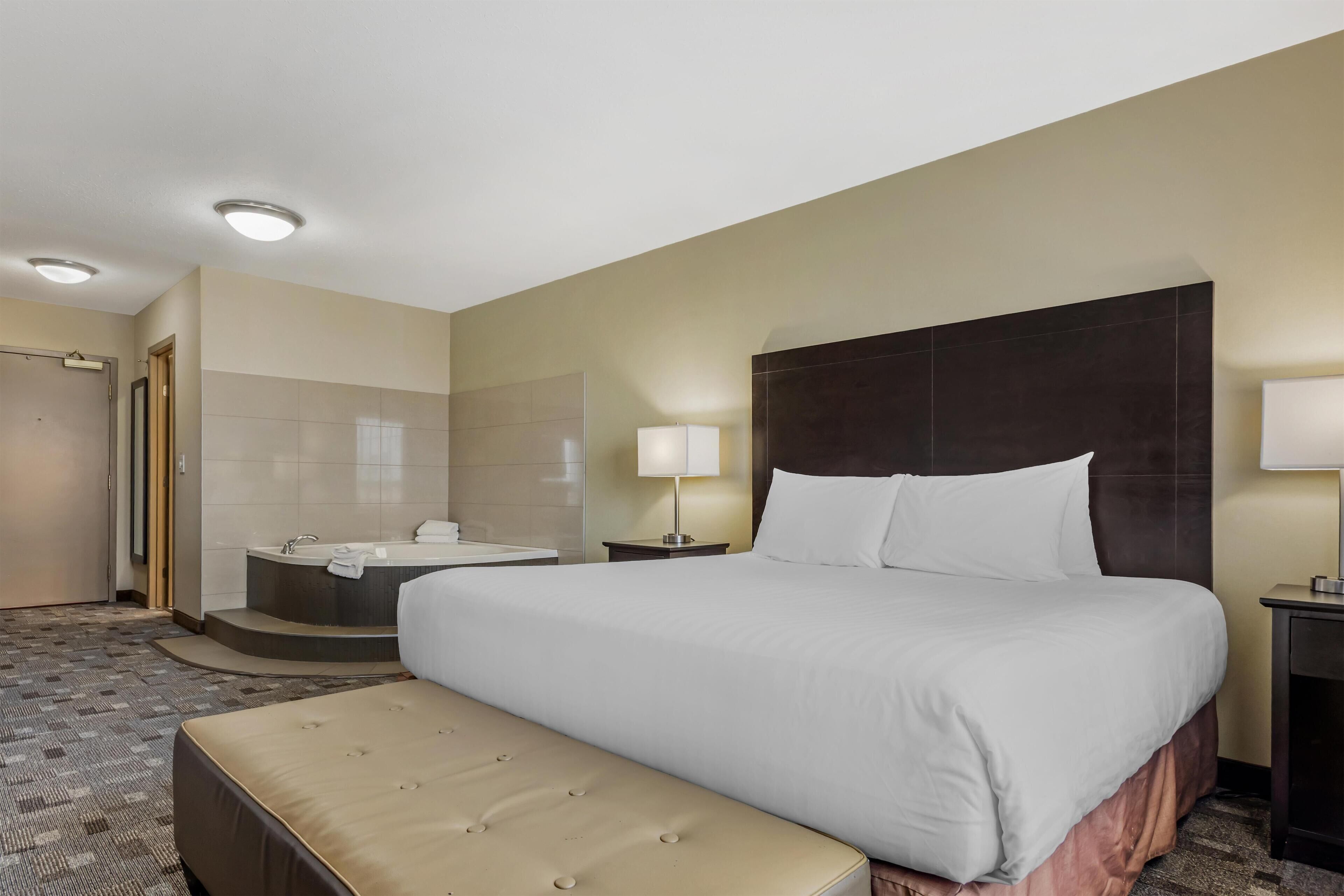A hotel room with a large bed, a separate bathtub, a small bedside table with a lamp, and a carpeted floor.