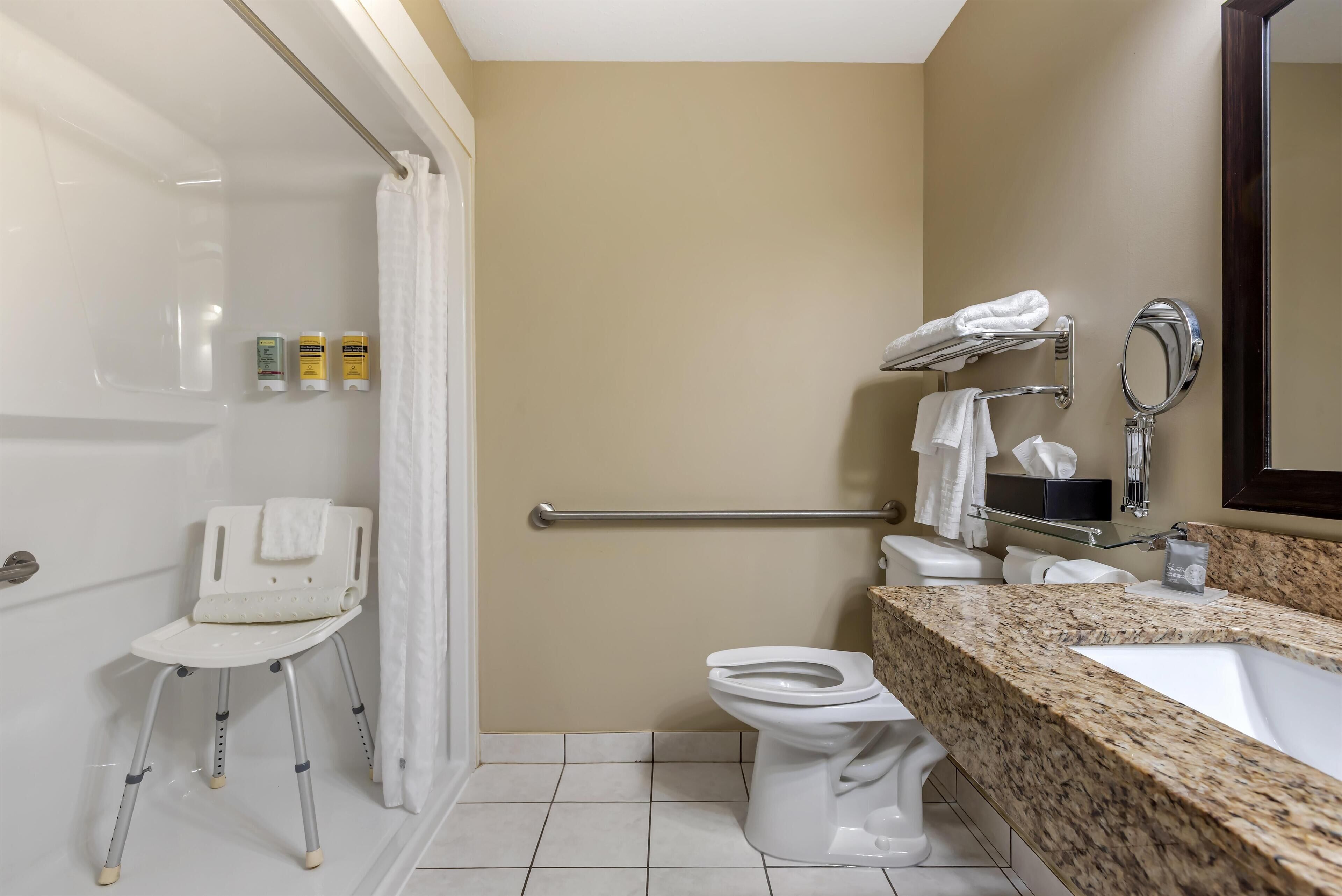 A bathroom with a shower chair, grab bars, a toilet, and a sink with a granite countertop.