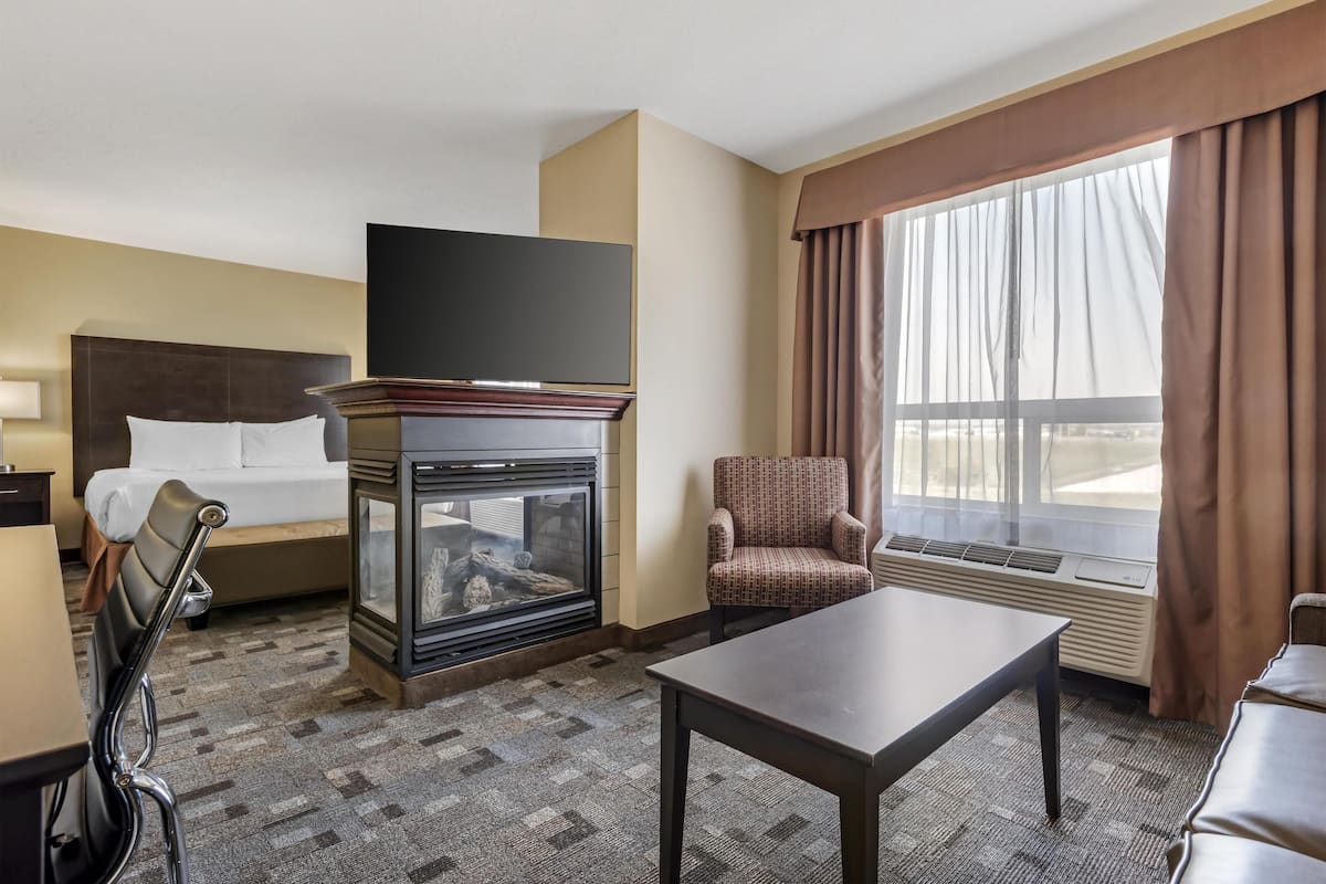 A hotel room with a bed, a flat-screen TV, a fireplace, a table, a chair, and a large window with curtains.