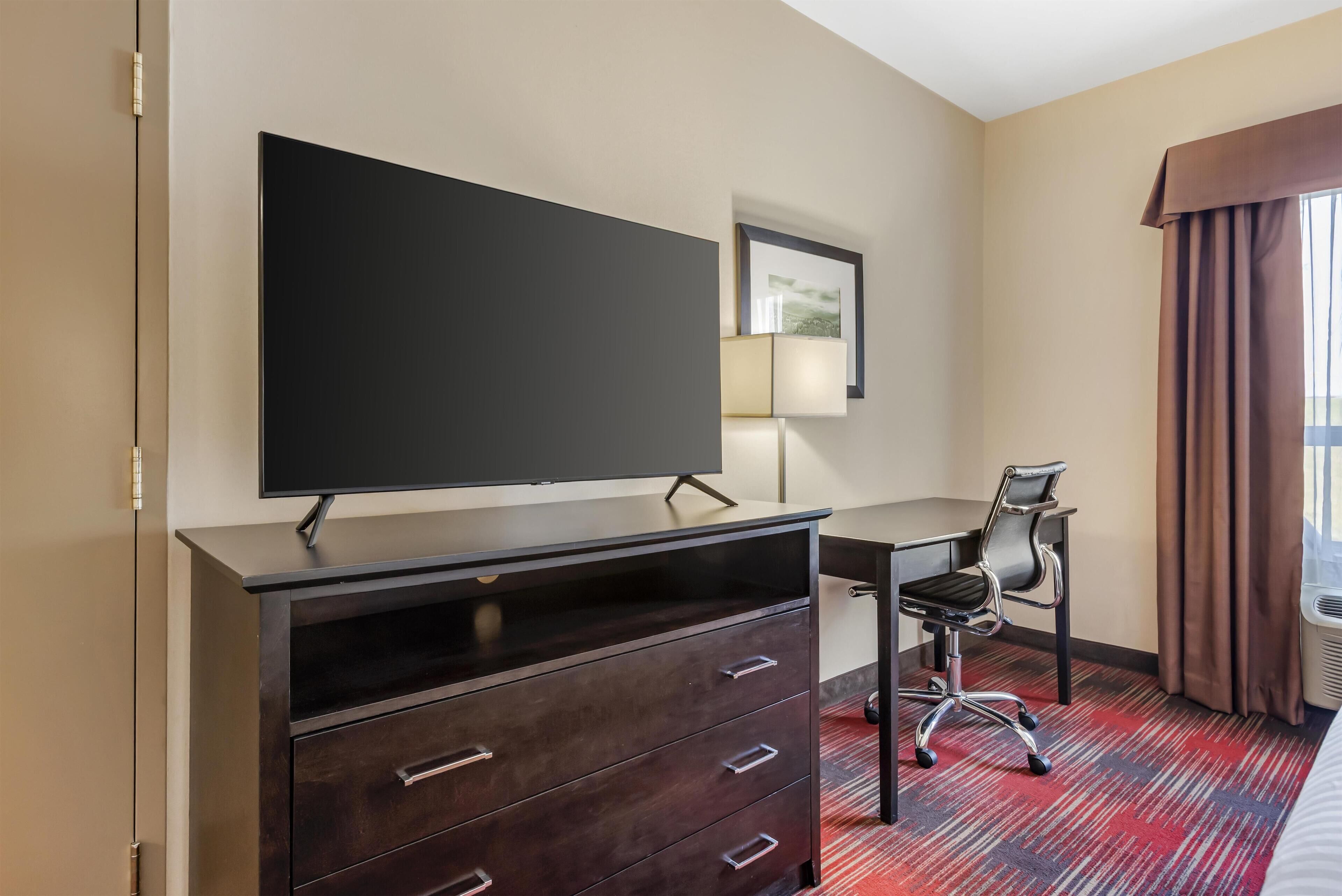 Habitación de hotel con televisor de pantalla plana sobre una cómoda de madera oscura, un escritorio con una silla, un cuadro en la pared y una ventana con cortinas.