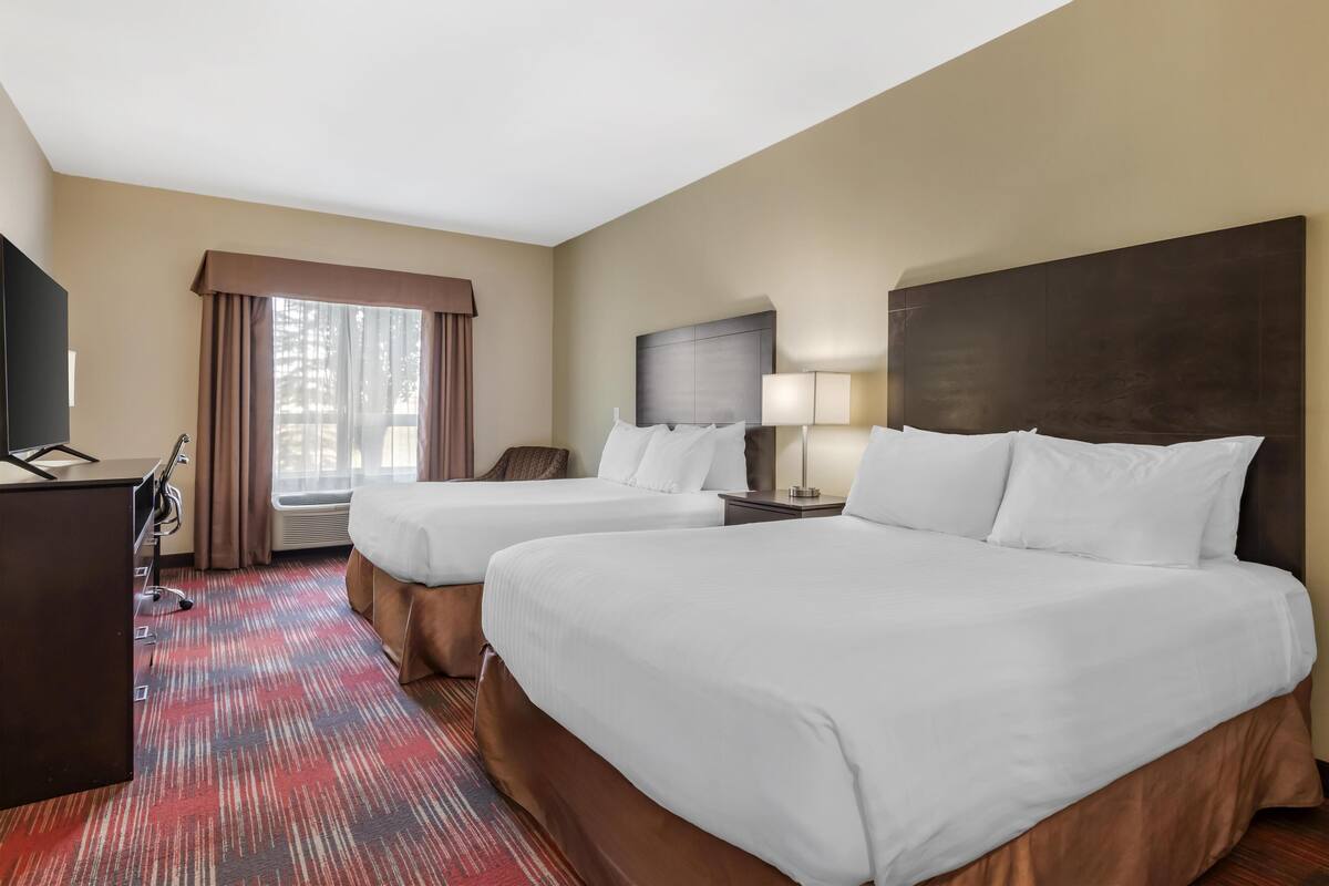 A hotel room with two beds, a desk, a lamp, and a large window with curtains.