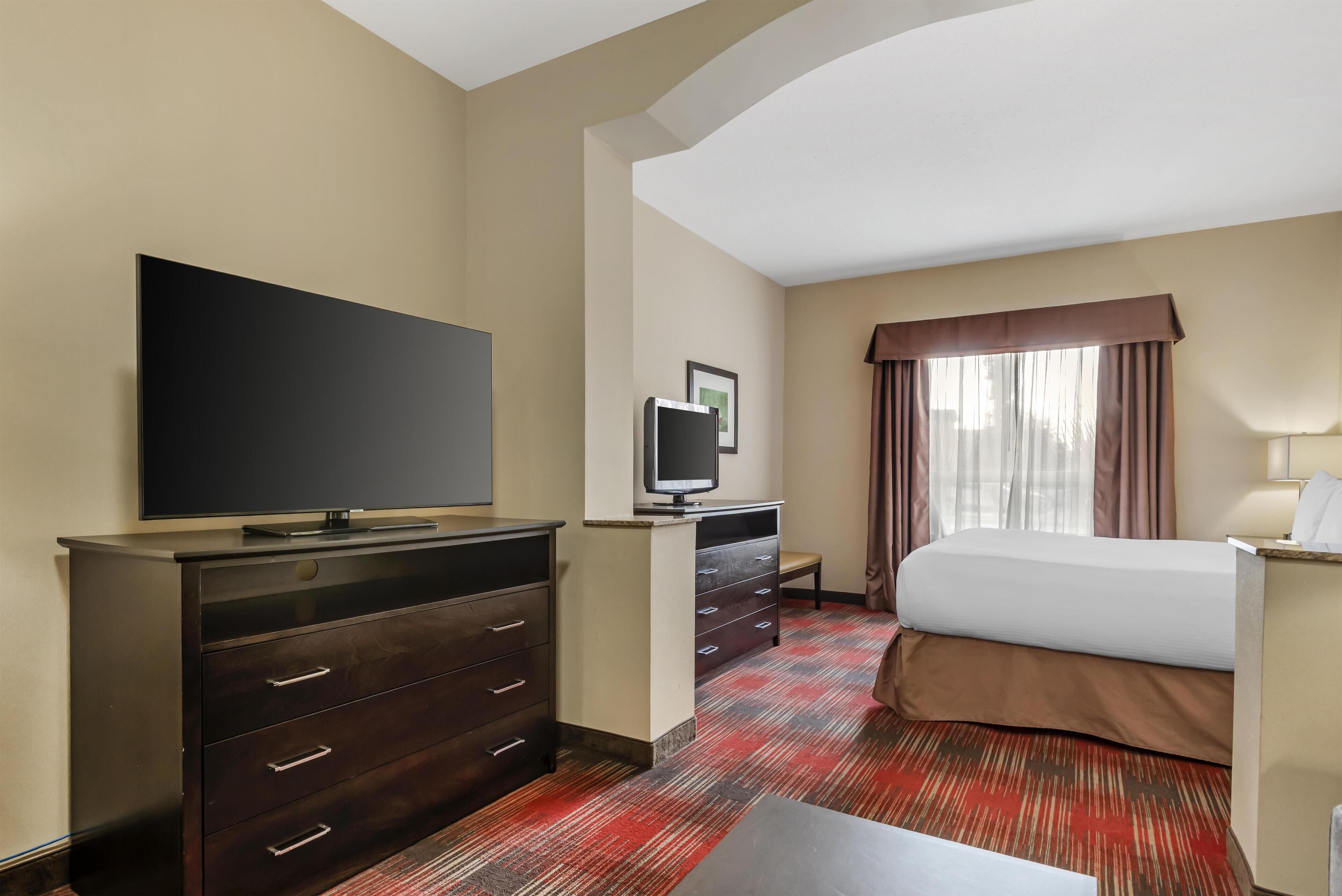 A hotel room with a large flat-screen TV, a desk with a computer, a bed with a brown bedspread, and a window with curtains.