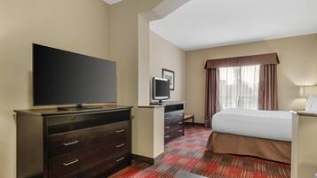 A hotel room with a large flat-screen TV, a desk with a computer, a bed with a brown bedspread, and a window with curtains.