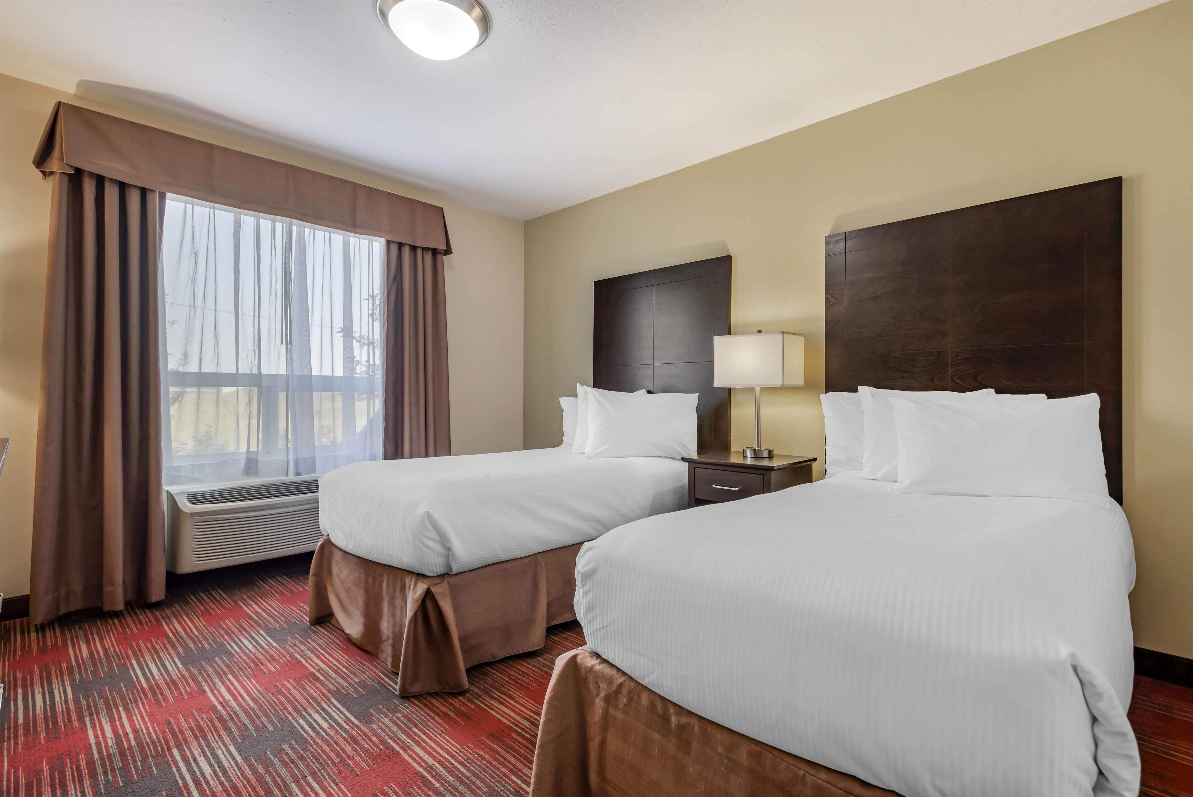 A hotel room with two beds, a window with curtains, a bedside table with a lamp, and a red carpet.