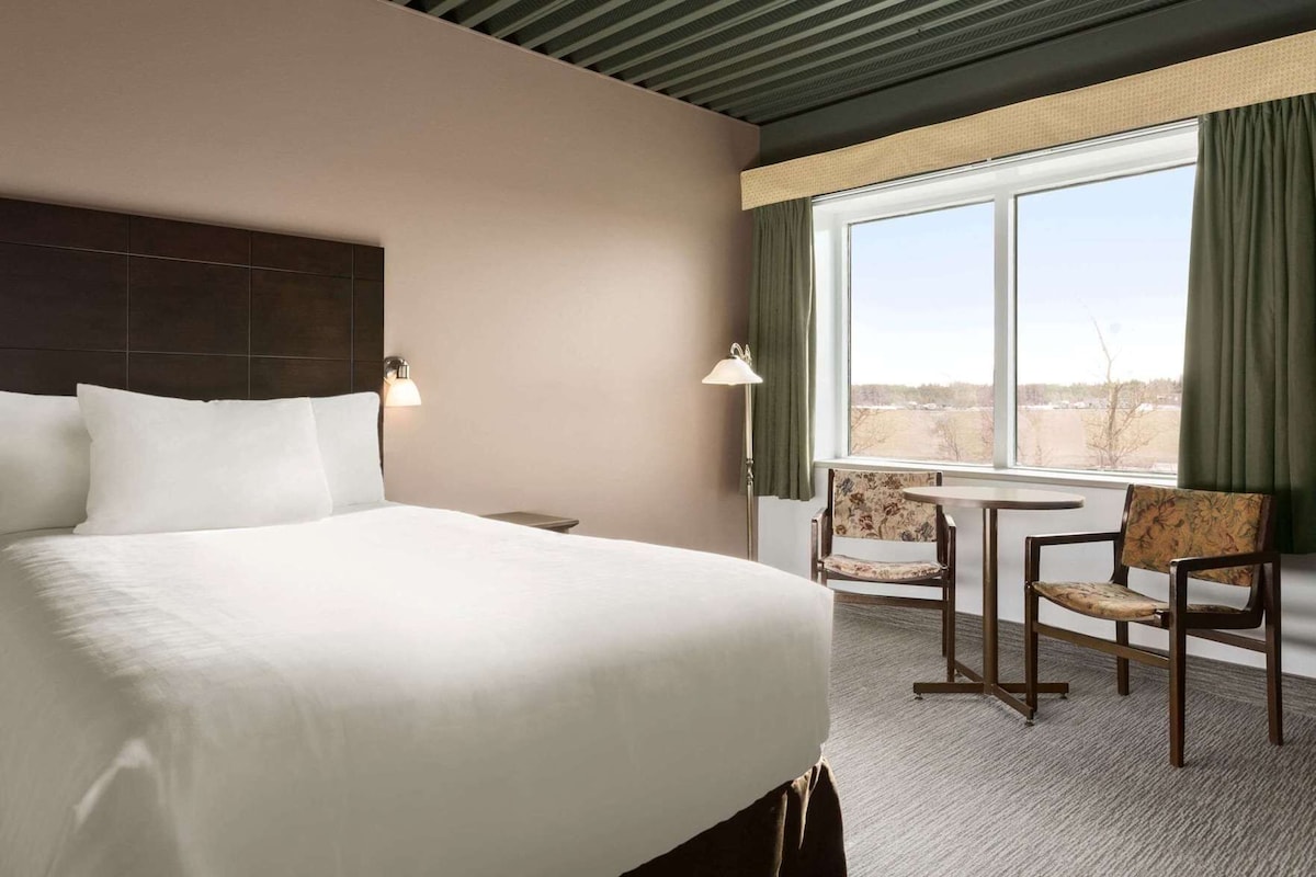 A hotel room with a large bed, a small table with two chairs, a window with curtains, and a lamp.